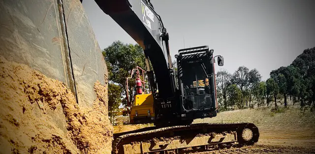 A large black excavator uses its arm to dig into a sandy embankment on a construction site, with trees and a clear sky visible in the background.