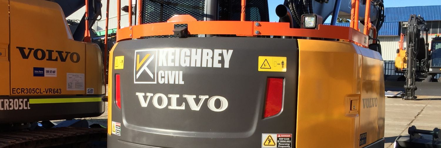 A close-up view of the back of a yellow Volvo construction vehicle labeled "Keighrey Civil," parked next to another Volvo vehicle on a sunny day at an industrial site.