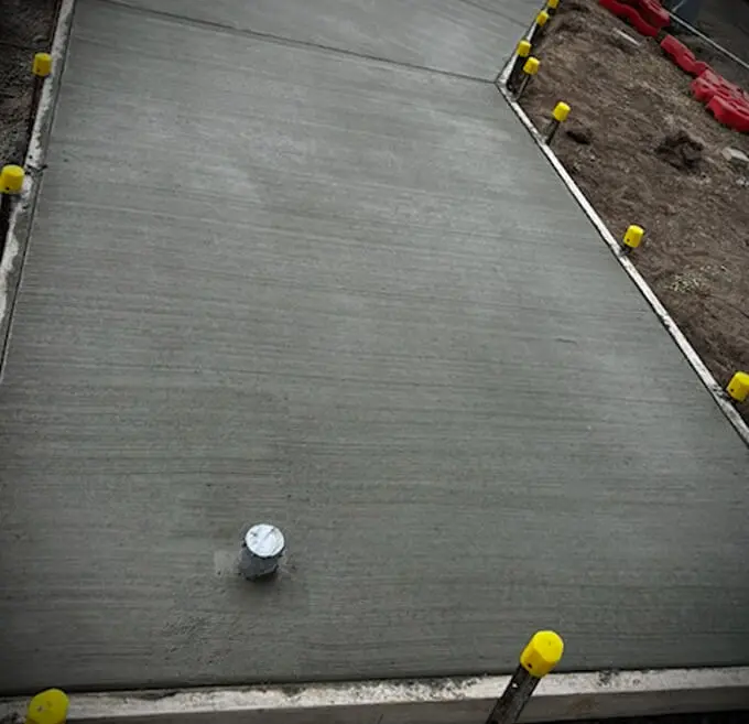 Freshly poured concrete sidewalk with a small pipe sticking up near the bottom left. Yellow caps cover the metal stakes along the edge, and dirt is visible on the right.