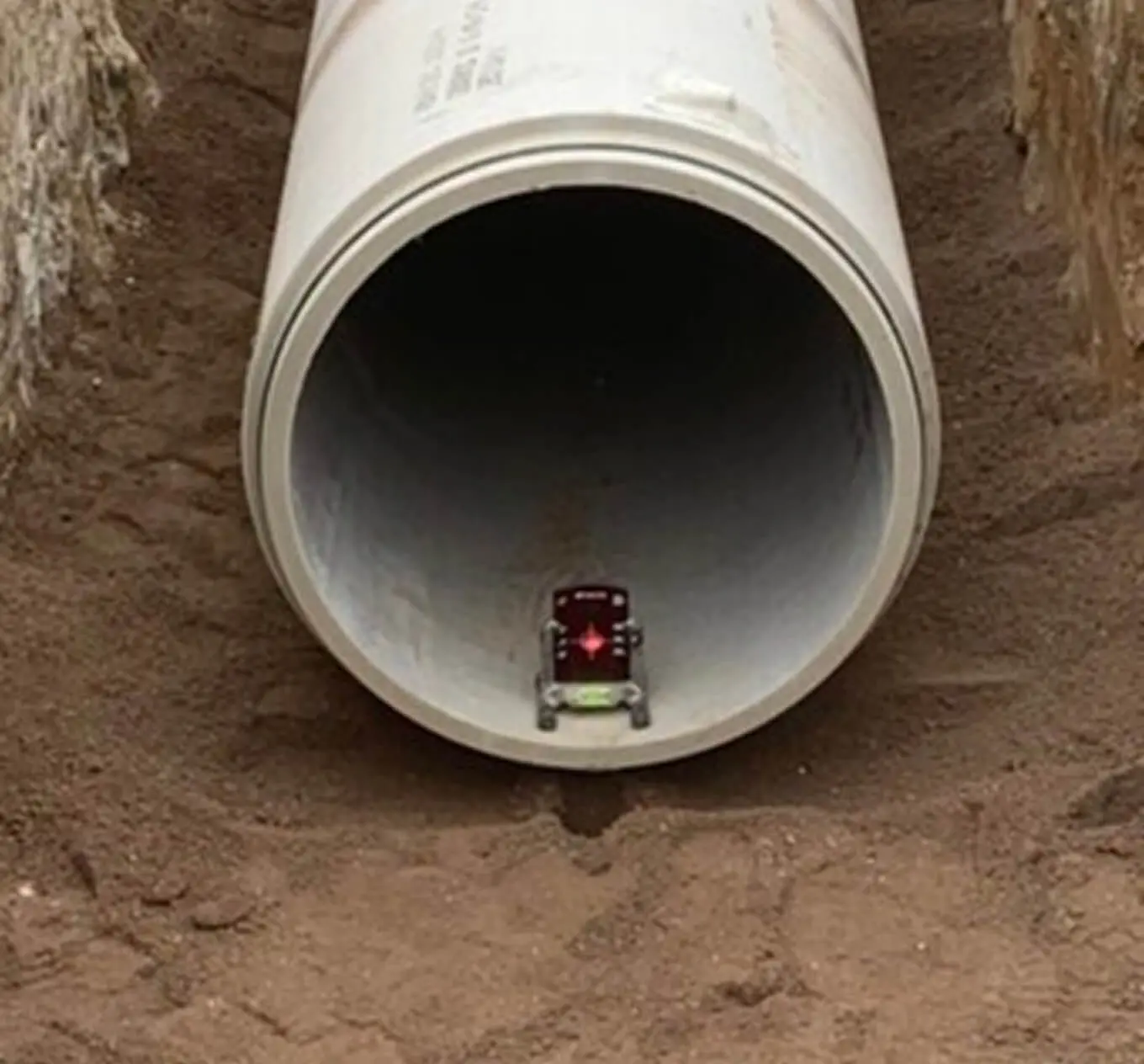 A toy car is positioned at the opening of a large concrete pipe lying horizontally in a dirt trench. The pipe makes the toy car appear very small by comparison.