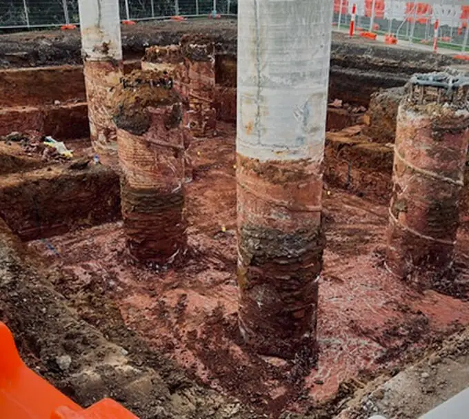 Archaeological excavation site with several large, circular brick and stone columns partially buried in soil, surrounded by orange safety barriers and fencing.