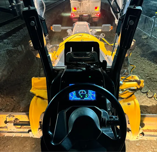 View from inside a construction vehicle cab, showing the steering wheel, digital dashboard, and controls, with a yellow arm and a truck visible outside on a work site at night.