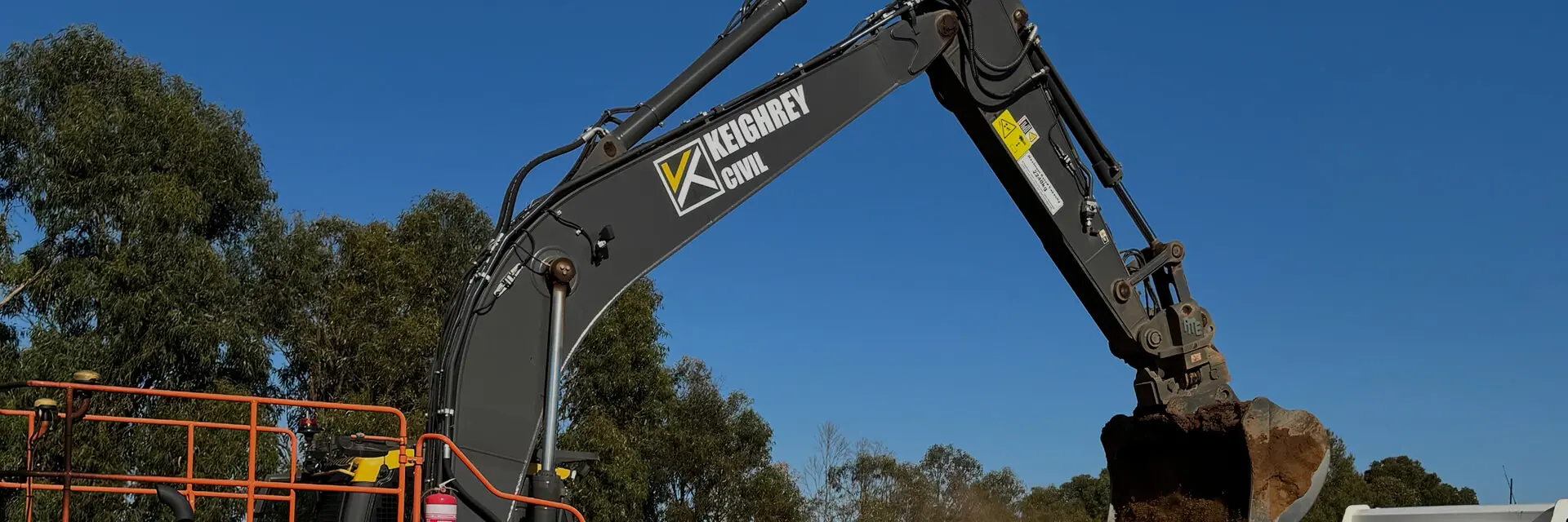 A large excavator with the logo "Keighray Civil" lifts a bucket of dirt at a construction site, with trees and a clear blue sky in the background.