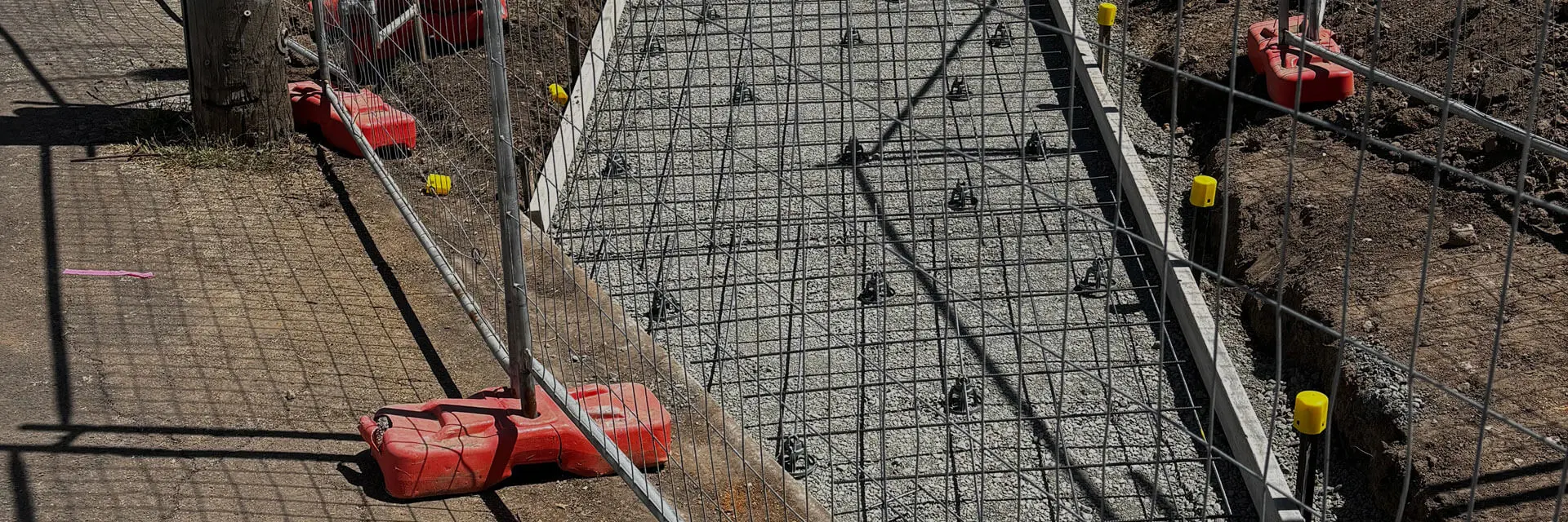 A construction site with a fenced-off area, showing a pathway covered in gravel and reinforcement mesh, possibly prepared for concrete. Red and yellow safety barriers line the edge; dirt and pavement are visible beside the site.
