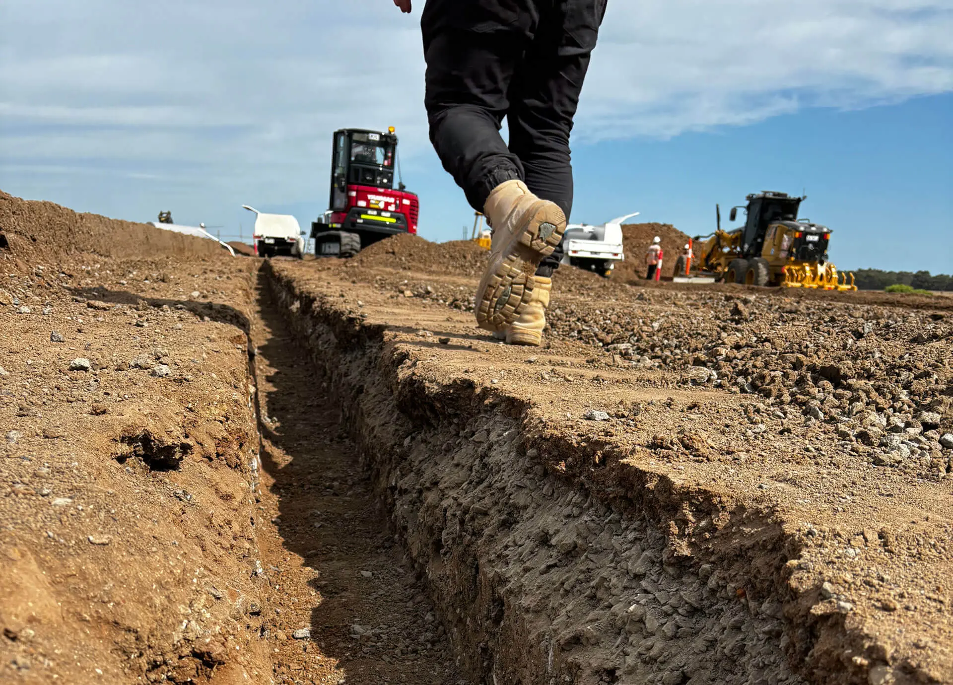 Close-up of a person in tan boots walking along a narrow dirt trench at a construction site, with machinery and workers visible in the background under a partly cloudy sky.