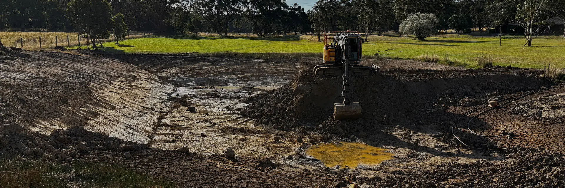 A large excavator is digging in a mostly dry, muddy pond surrounded by trees and grass on a sunny day. The pond's water level is very low, leaving exposed earth and patches of mud.