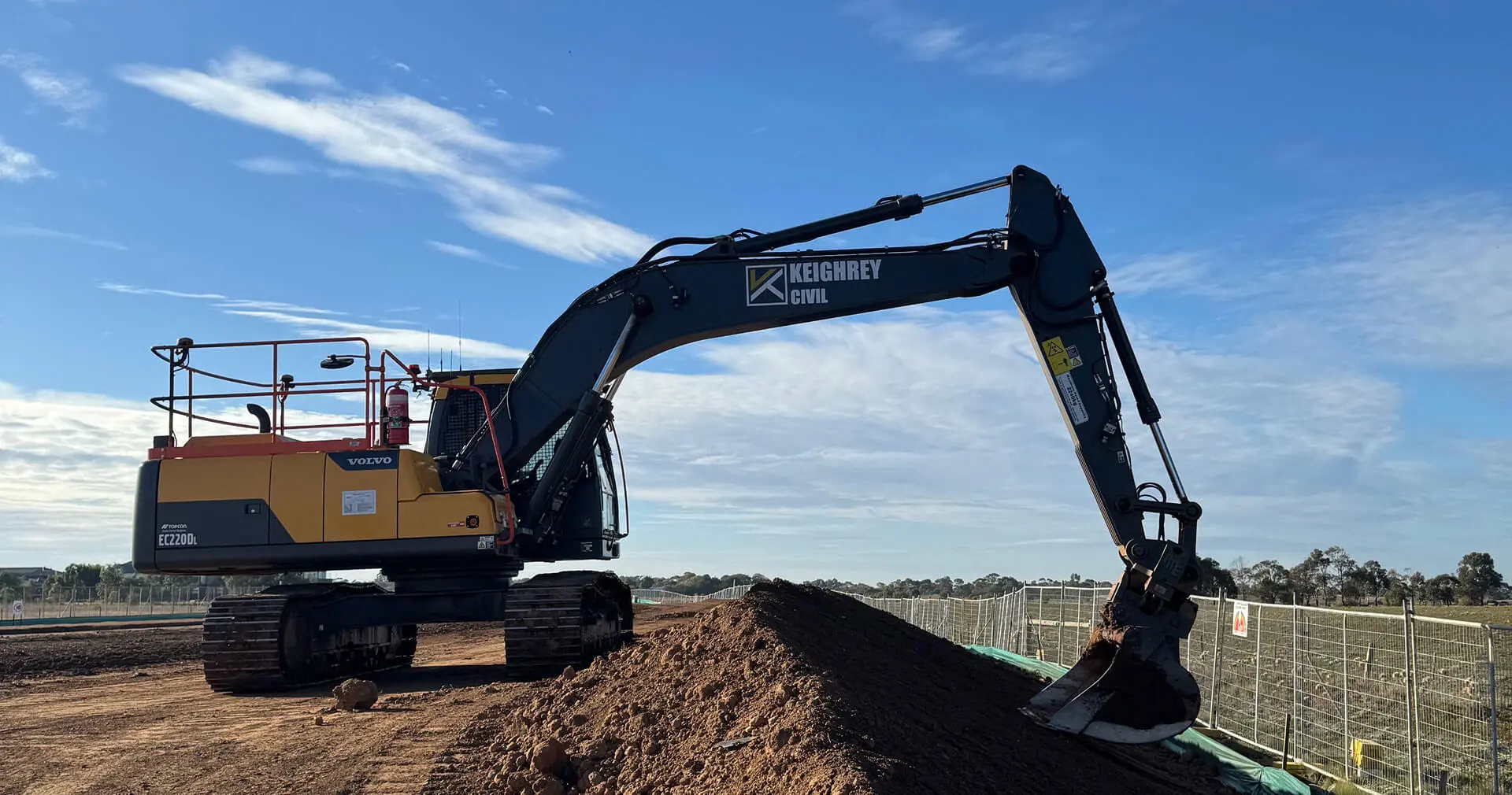 A large excavator is digging into a mound of dirt at a construction site under a blue sky with clouds. Fencing and open land are visible in the background.